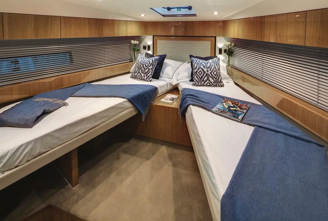 A stylish boat cabin features two single beds with blue throws, decorative cushions, and a magazine on the floor.
